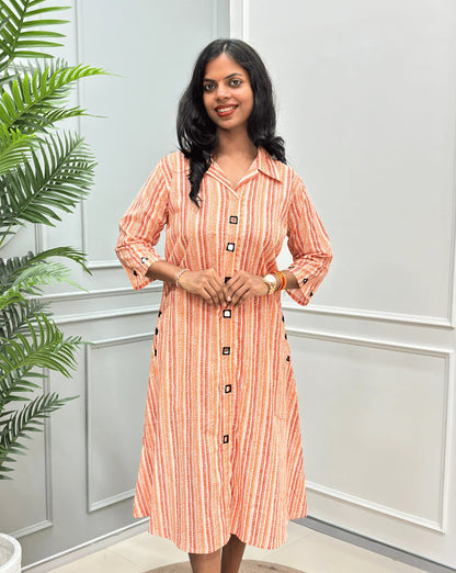 Front-View of Rust Orange Women's Ajrakh A-line Pure Cotton Long Kurti with an Ajrakh Strip Print on abstract of orange, white, Rust orange, Real hand-mirror work on front & Sleeves, Along with 2 side pockets, standing on a round woven rug in front of a light gray paneled wall with a potted plant.