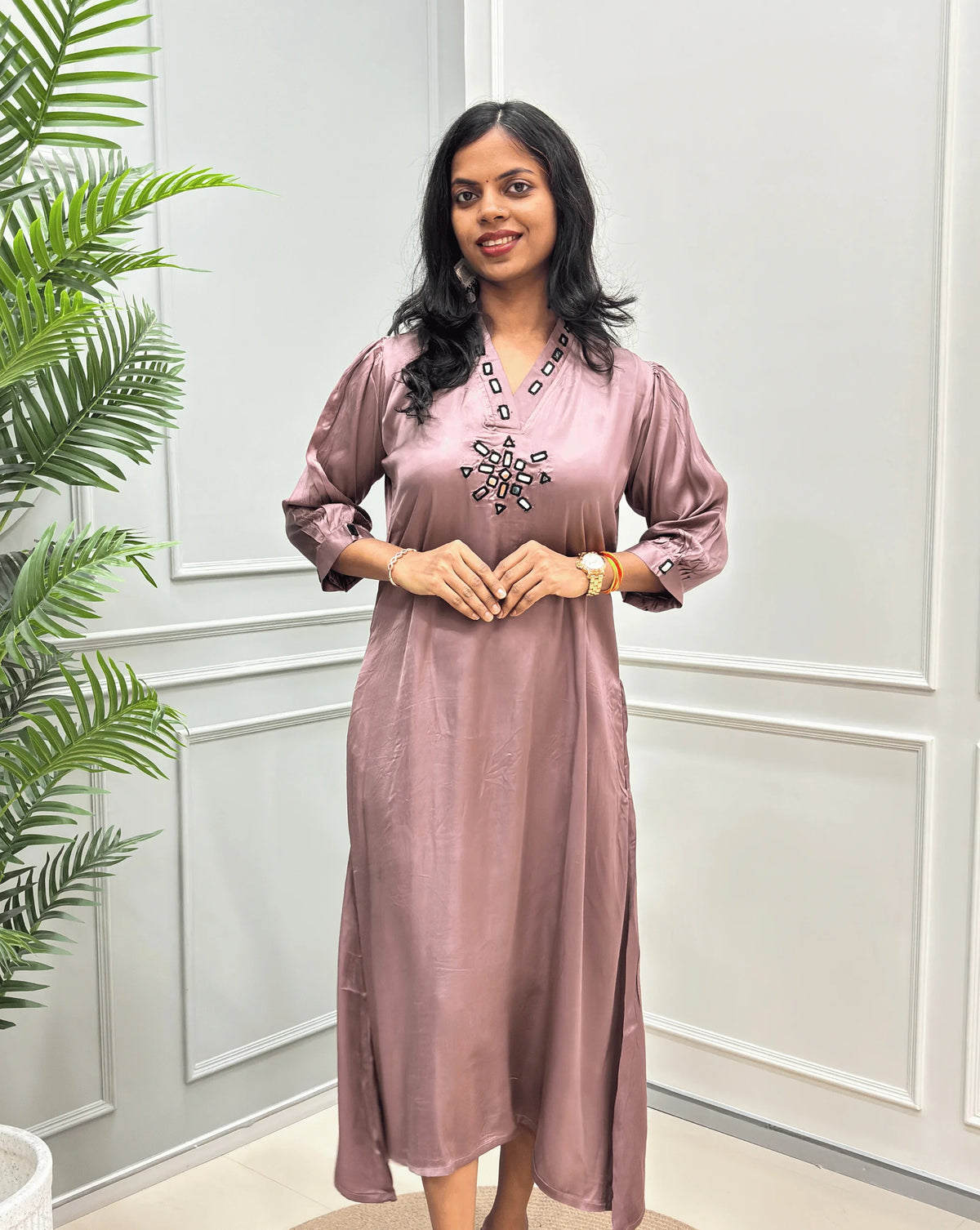 Front -View of Onion Pink Women's Pure Modal Silk Ajrakh Long Kurti with a decorative Real Hand-Mirror embroidered V-neckline, Floral pattern on front & Sleeves Cuffs, standing on a round woven rug in front of a white paneled wall with a potted plant on the left