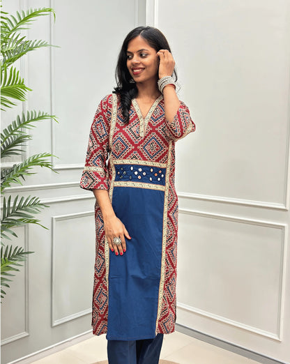 Front View of Blue Women's Ajrakh pure cotton 2 peice Co-Ord set with a Red base & an abstract of blue & Beige Bandhani print on Front Chest to Side & Sleeves, Hand-mirror work on Front as Belt, standing on a round woven mat in front of a white paneled wall with a potted plant.