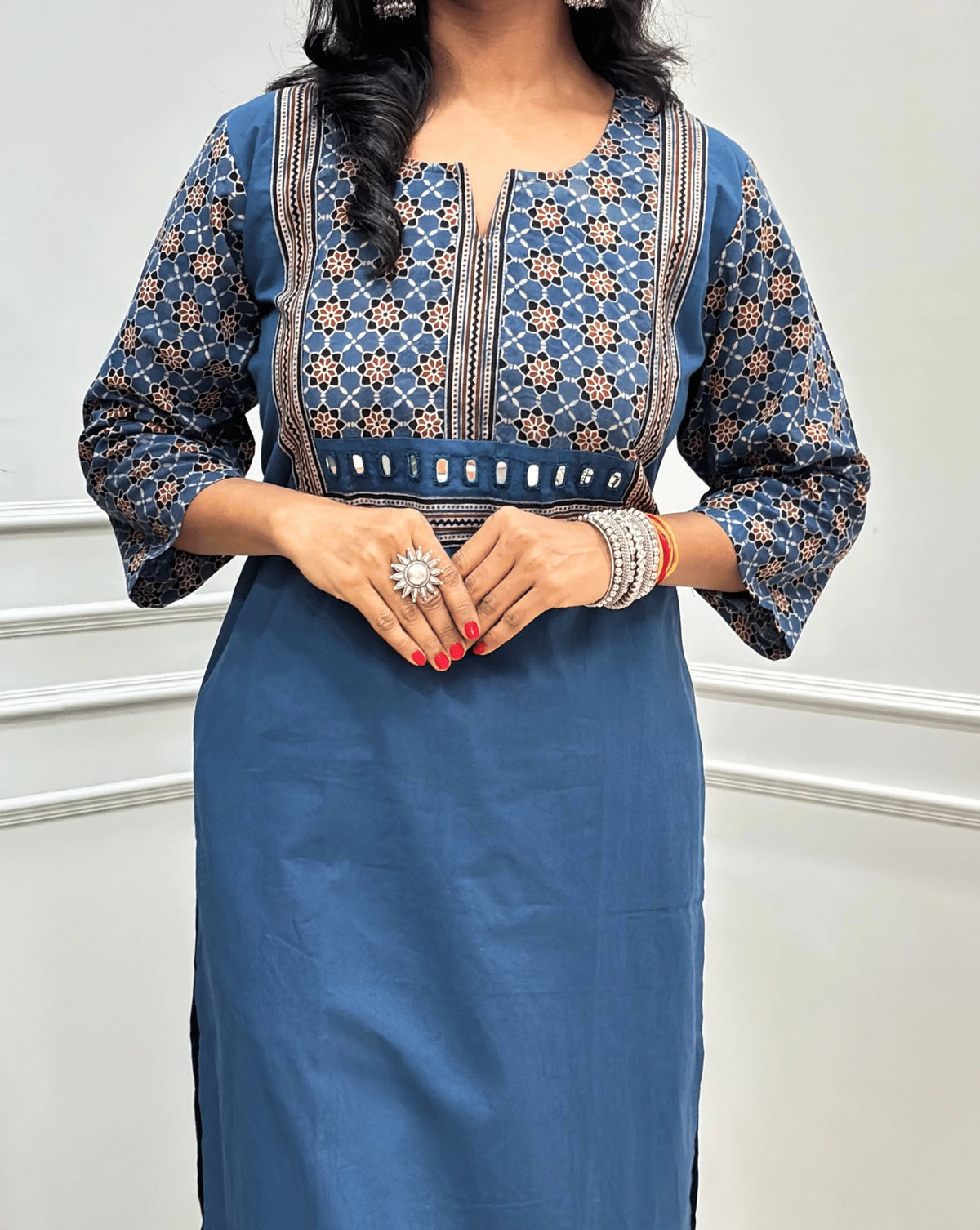 Close-up view of Light Blue Women's Hand-block Ajrakh Pure Cotton Long Kurti featured with Round V Shaped neck along with three quarter sleeves, with a blue base, an abstract of black & beige Floral Print yoke on front & Full sleeves, Hand-mirror work on front, paired with blue Pant, standing on a round woven mat in front of a white paneled wall. 