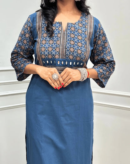 Close-up view of Light Blue Women's Hand-block Ajrakh Pure Cotton Long Kurti featured with Round V Shaped neck along with three quarter sleeves, with a blue base, an abstract of black & beige Floral Print yoke on front & Full sleeves, Hand-mirror work on front, paired with blue Pant, standing on a round woven mat in front of a white paneled wall. 