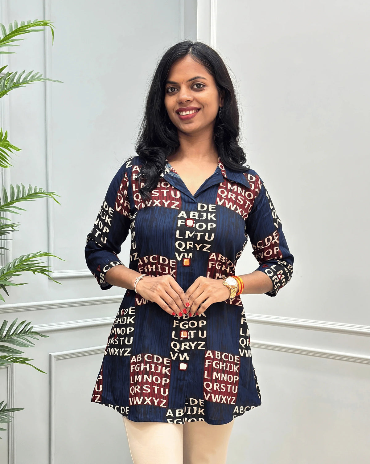 Front View of Navy Blue Women's Ajrakh Vegetable Dyed Plant based Cotton Short kurti with a Red & Black Base, & Abstract Beige color alphabet print, Hand-mirror work on Front, Sleeves & Pockets paired with beige trousers, standing on a round woven rug in front of a white paneled wall with a potted plant.
