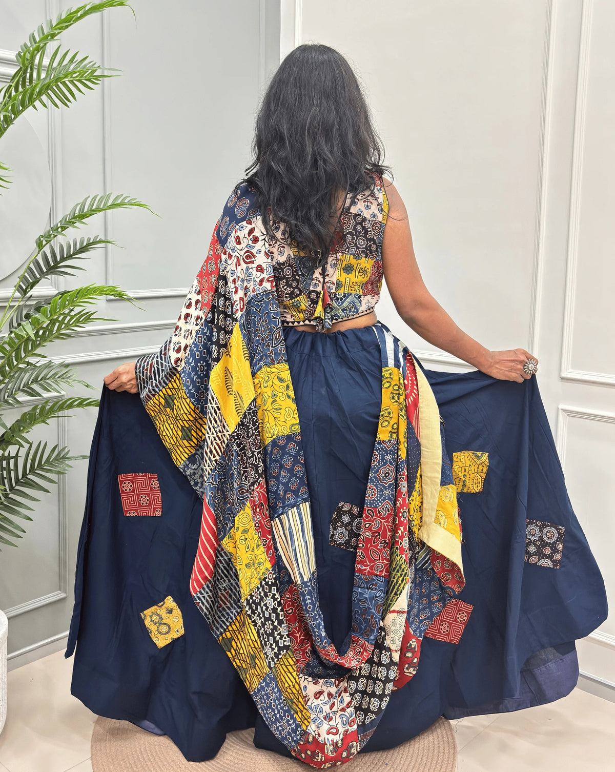 Back-Side View of Navy blue Women's Ajrakh pure Cotton Skirt Set with a Ajrakh Patch work along with multi-colour Printed blouse & Dupatta, Standing against a light paneled wall with a green plant on the left.