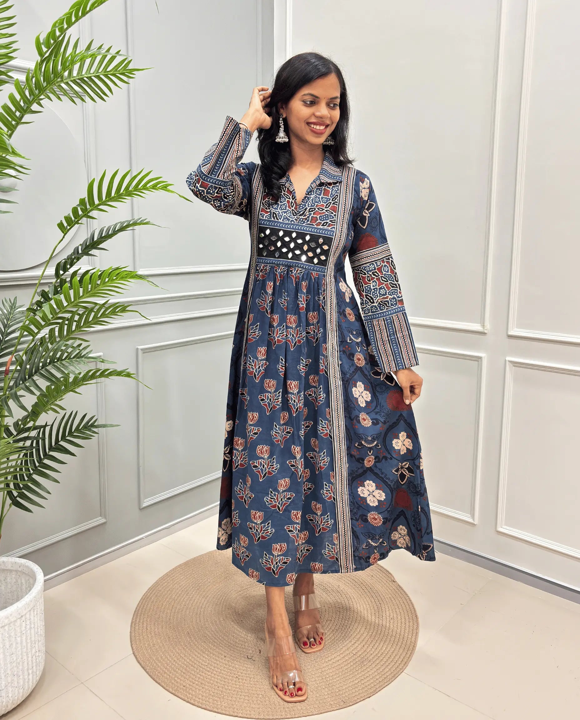                                                                                                 
Blue Women's Cotton Hand-block Long kurti with intricate floral and geometric block prints in beige, white, and red. The dress features a V-neckline, a patterned waistband, and a gathered skirt, shown in a styled indoor setting with a potted plant and paneled walls.