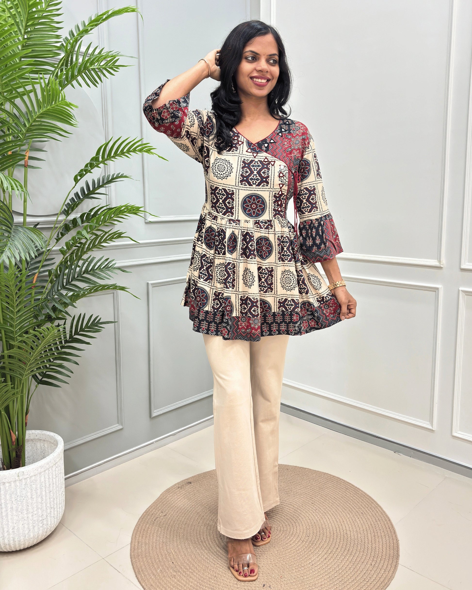 Beige Women's Ajrakh Vegetable Dyed plant based cotton Short Kurti with a black and red geometric pattern, paired with beige flared pants, standing on a round woven mat in front of a white paneled wall with a potted plant.