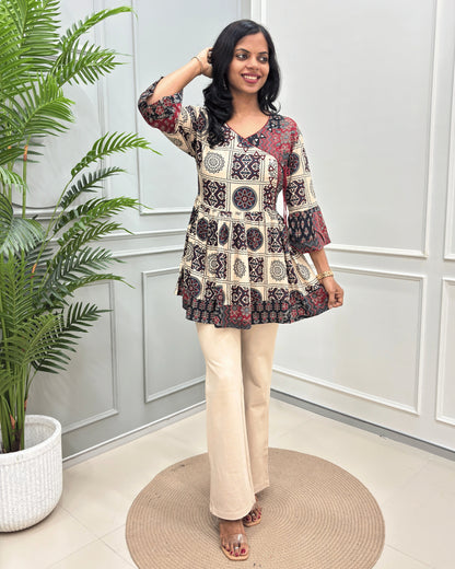 Beige Women's Ajrakh Vegetable Dyed plant based cotton Short Kurti with a black and red geometric pattern, paired with beige flared pants, standing on a round woven mat in front of a white paneled wall with a potted plant.