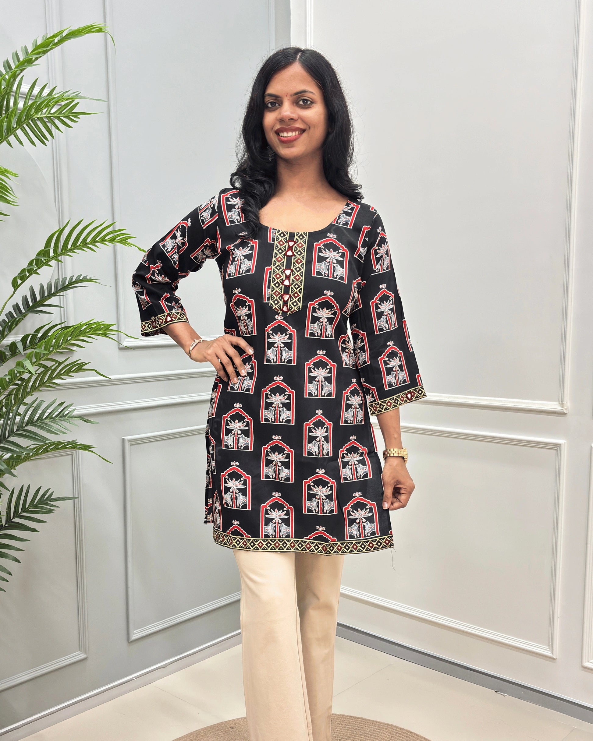 Front view of Black Women's Ajrakh plant based Cotton Short Kurti with a red and Grey Pichhwai pattern, paired with beige trousers, standing in front of a white paneled wall with a green plant on the left.