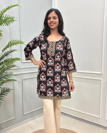 Front view of Black Women's Ajrakh plant based Cotton Short Kurti with a red and Grey Pichhwai pattern, paired with beige trousers, standing in front of a white paneled wall with a green plant on the left.