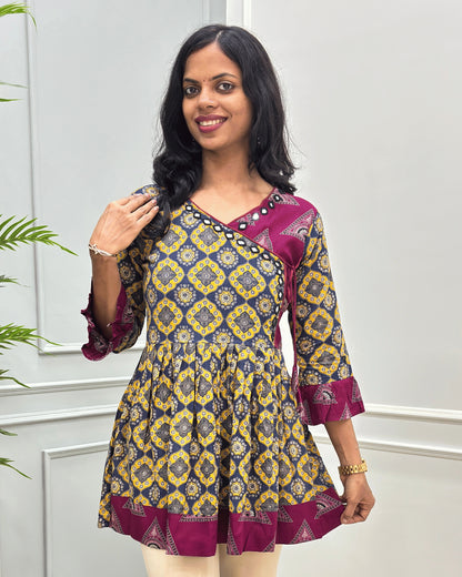 Close-up of Yellow plant based cotton  short kurti with three-quarter ruffled sleeves, beautiful Angrakha pattern with lace, hand-mirror work on V-neck standing against a light gray paneled wall