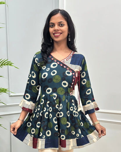 Close-up of Green women's Ajrakh plant based cotton Short kurti with a colorful geometric and circular print, Hand-Mirror work on V- neckline along with lace, paired with beige pants, standing in front of a white paneled wall with a green plant on the left.