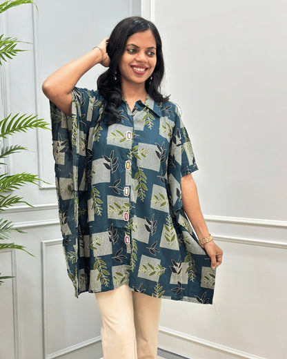 Front-view of Green Women's Ajrakh Mashru Silk short-sleeve button-up Kaftan top with a beige, and green botanical patchwork pattern, paired with beige pants, standing in front of a white paneled wall.