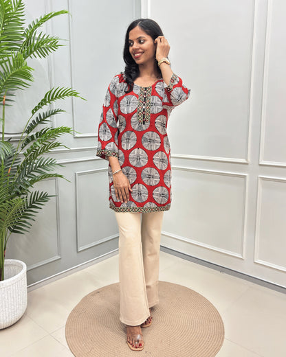 Red women's Vegetable dyed plant based Cotton Short kurti with a colorful geometric circular print and beige border trim, paired with beige wide-leg trousers, standing on a round woven mat in front of a white paneled wall.