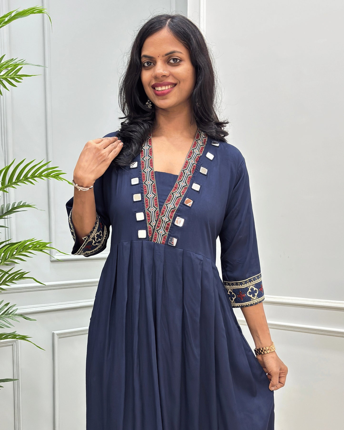 Close-up of navy blue women's plant based cotton long-kurti with colorful embroidered details on the neckline and cuffs,2 side pockets with hand-mirror work on border of pockets and neck outline, standing in front of a light gray paneled wall with a green plant on the left.