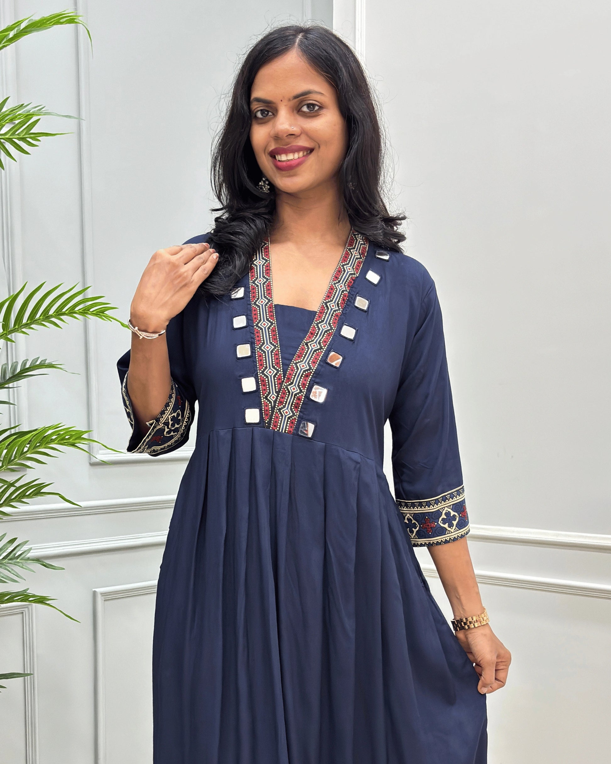 Close-up of navy blue women's plant based cotton long-kurti with colorful embroidered details on the neckline and cuffs,2 side pockets with hand-mirror work on border of pockets and neck outline, standing in front of a light gray paneled wall with a green plant on the left.