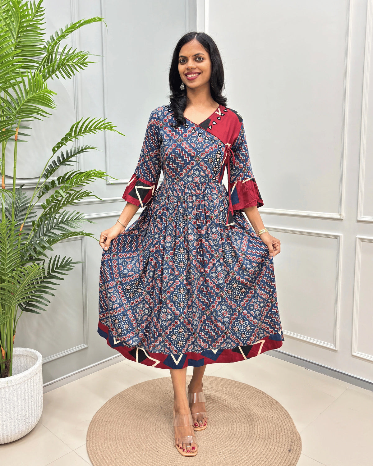 Blue women's Vegetable dyed cotton Long-Kurti with 3/4th Frill sleeves with geometric print on Red Base Colour on sleeves & bottom, Standing in front of white wall, left side Green plant .