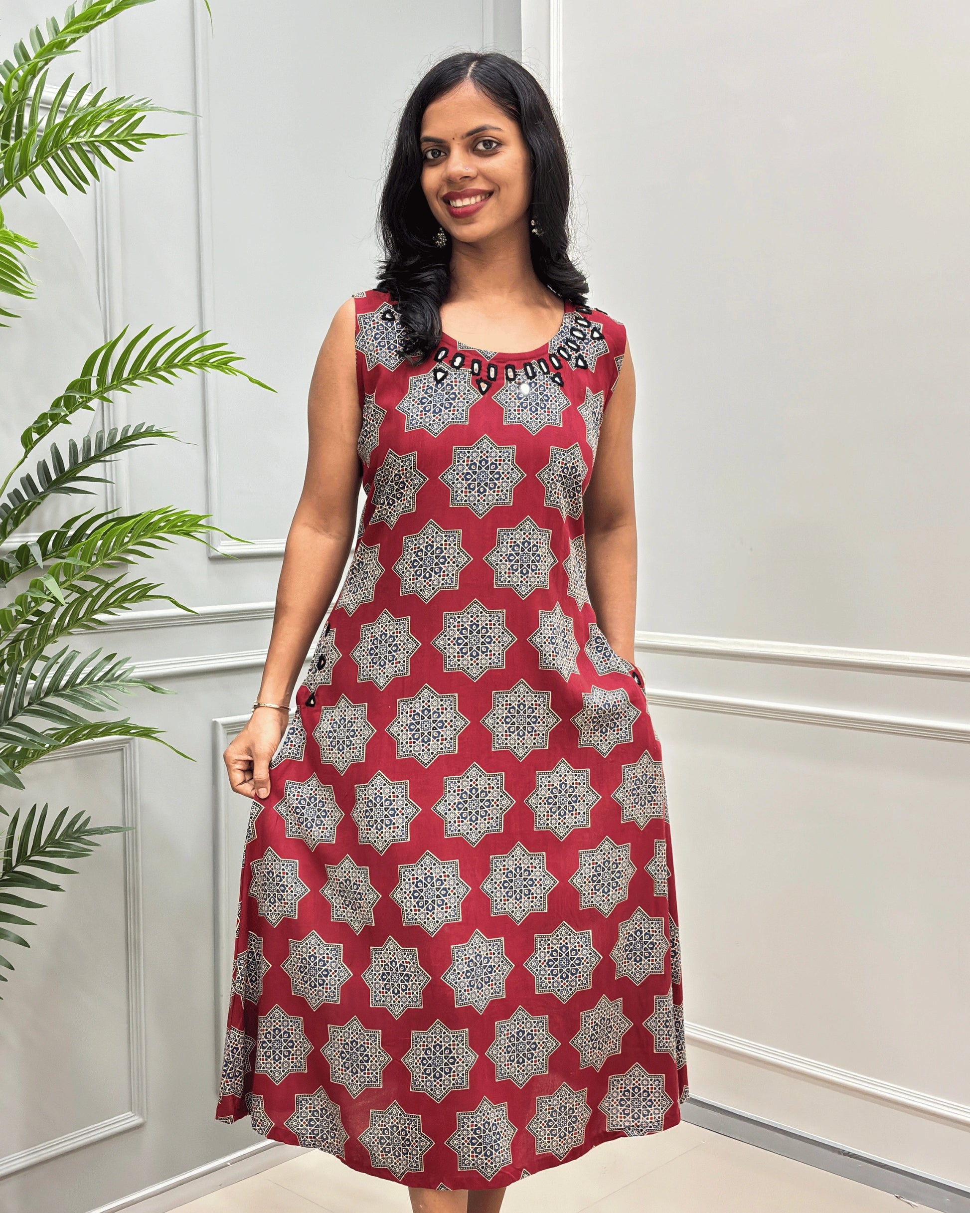 Red Women's plant based cotton Long sleeveless Kurti with a Beige and Black geometric star pattern and a black beaded on Hand-mirror neckline, standing in front of a white paneled wall with a green plant on the left.