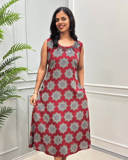Red Women's plant based cotton Long sleeveless Kurti with a Beige and Black geometric star pattern and a black beaded on Hand-mirror neckline, standing in front of a white paneled wall with a green plant on the left.