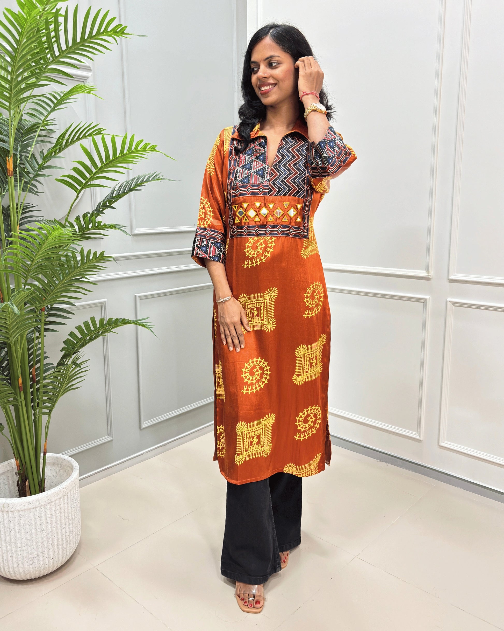 Orange women's Pure Mashru Silk Long Kurti with red , Black & white geometric patterns on the yoke and sleeves, paired with black Flared pants, standing in a room with a potted plant.