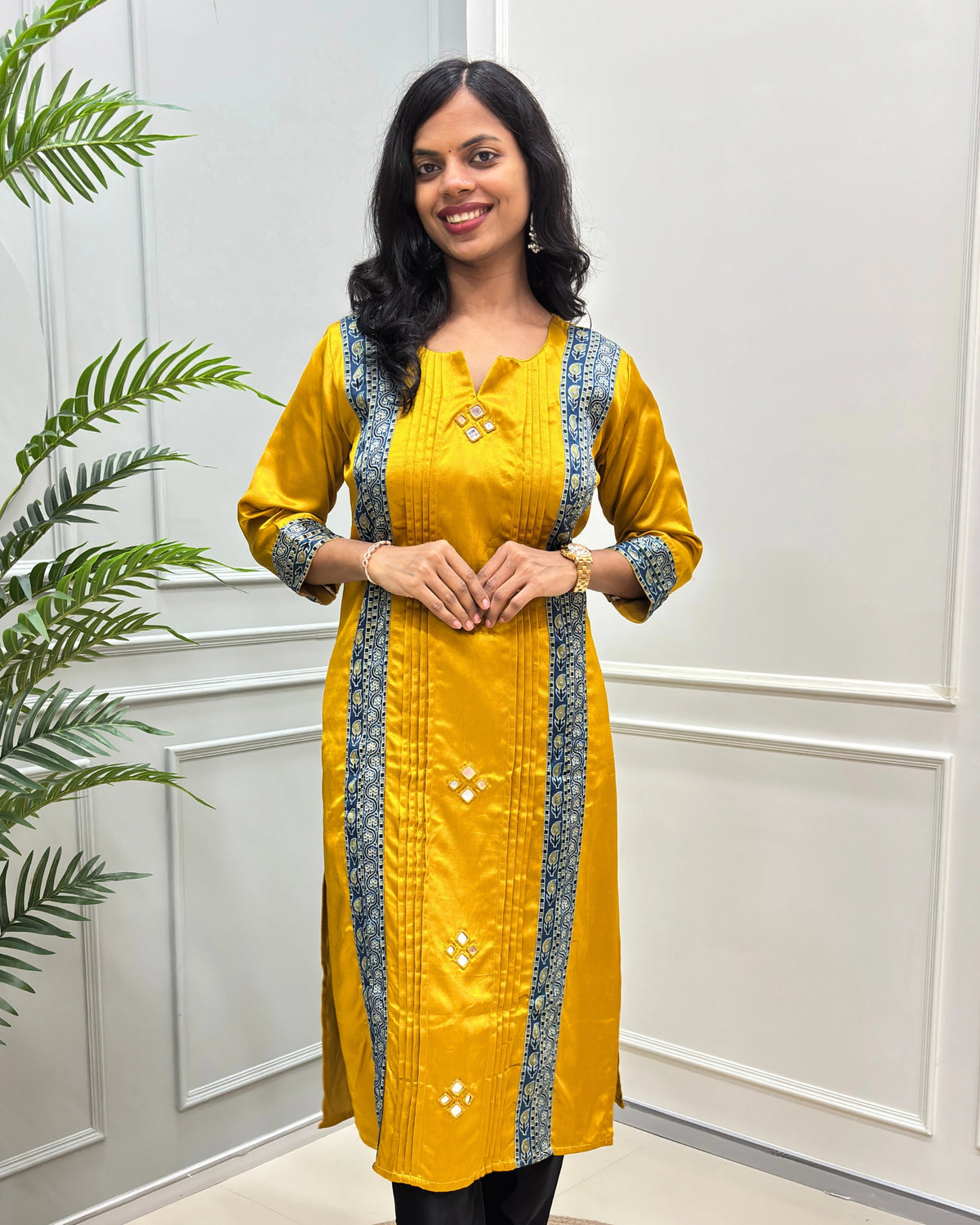 Front view of Yellow Ajrakh Mashru Silk Long-kurti with blue-Beige patterned vertical panels and matching patterned cuffs, standing in front of a white paneled wall with a green plant on the left.
