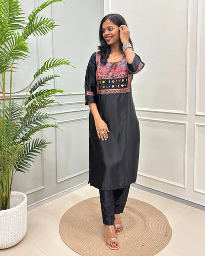 Black Women's Ajrakh Mahru Silk Long-kurti with a geometric Print yoke in blue, red, and white, with Hand-Mirror Work,  paired with black pants, standing on a round woven mat in front of a white paneled wall with a potted plant.