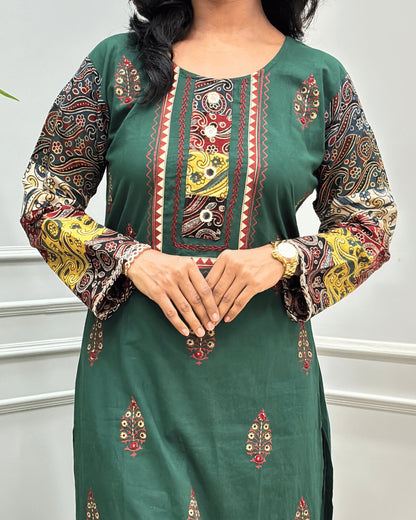 Close-up of Green Rayon Cotton Co-Ord Set with intricate multicolor printed patterns on Full-sleeves, neckline with block , and Hand-mirror motifs on the front, standing against a light gray paneled wall.