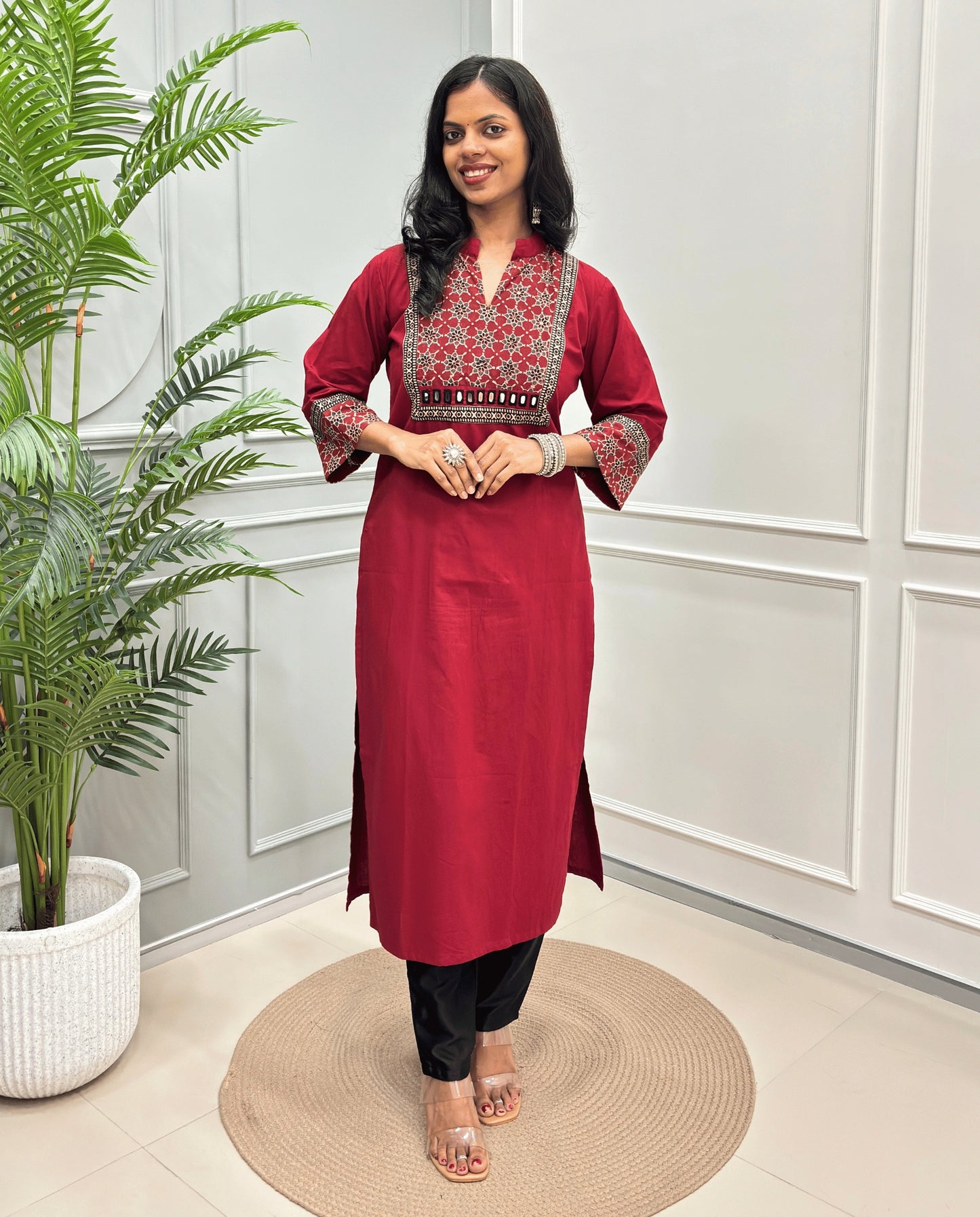 Red Women's Hand-block Ajrakh Pure Cotton Long kurti with intricate Beige & black geometric print on the chest and sleeve cuffs, paired with black pants , standing on a round woven mat in front of a white paneled wall with a potted plant.