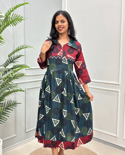 Bottle-Green Women's Ajrakh Vegetable dyed plant based Cotton Long-Kurti with a Red neck and cuffs, and a Green-colored collar & dress featuring a geometric pattern of triangles and spiral in beige, Green, and blue, Hand-mirror work on front, standing on a round woven rug in front of a white paneled wall with a green potted plant on the left.