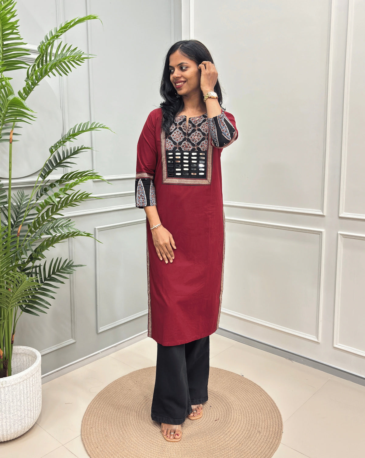 Red Women's Hand-block Ajrakh Pure Cotton Long Kurti with a red base and abstract Black, Red & Beige  geometric embroidery on the chest and sleeve cuffs, paired with black Flared pants, standing in a decorated room with a potted plant.
