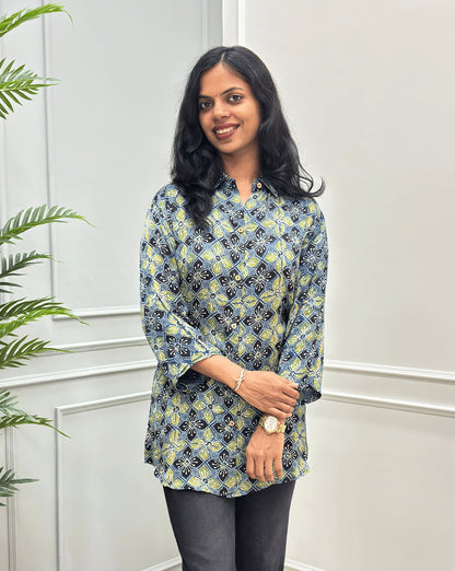 Front View of Green Women's Ajrakh Pure Modal Silk Shirt with a Floral print on blue, yellow, and white geometric pattern, black flared pants, standing on a round woven rug in front of a white paneled wall with a potted plant on the left