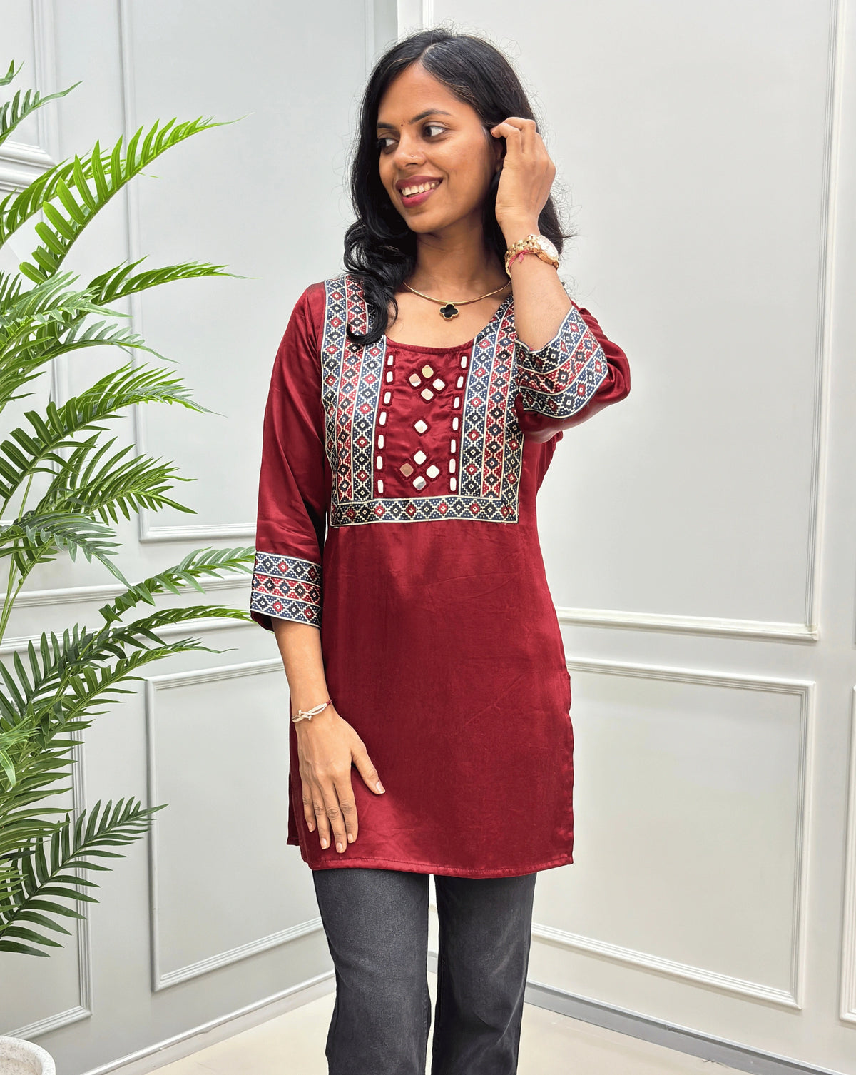 Close-up of mashru silk Red short kurti  with intricate multicolor geometric embroidery on the neckline and sleeves, paired with dark gray pants, standing in front of a white paneled wall with a green plant on the left.