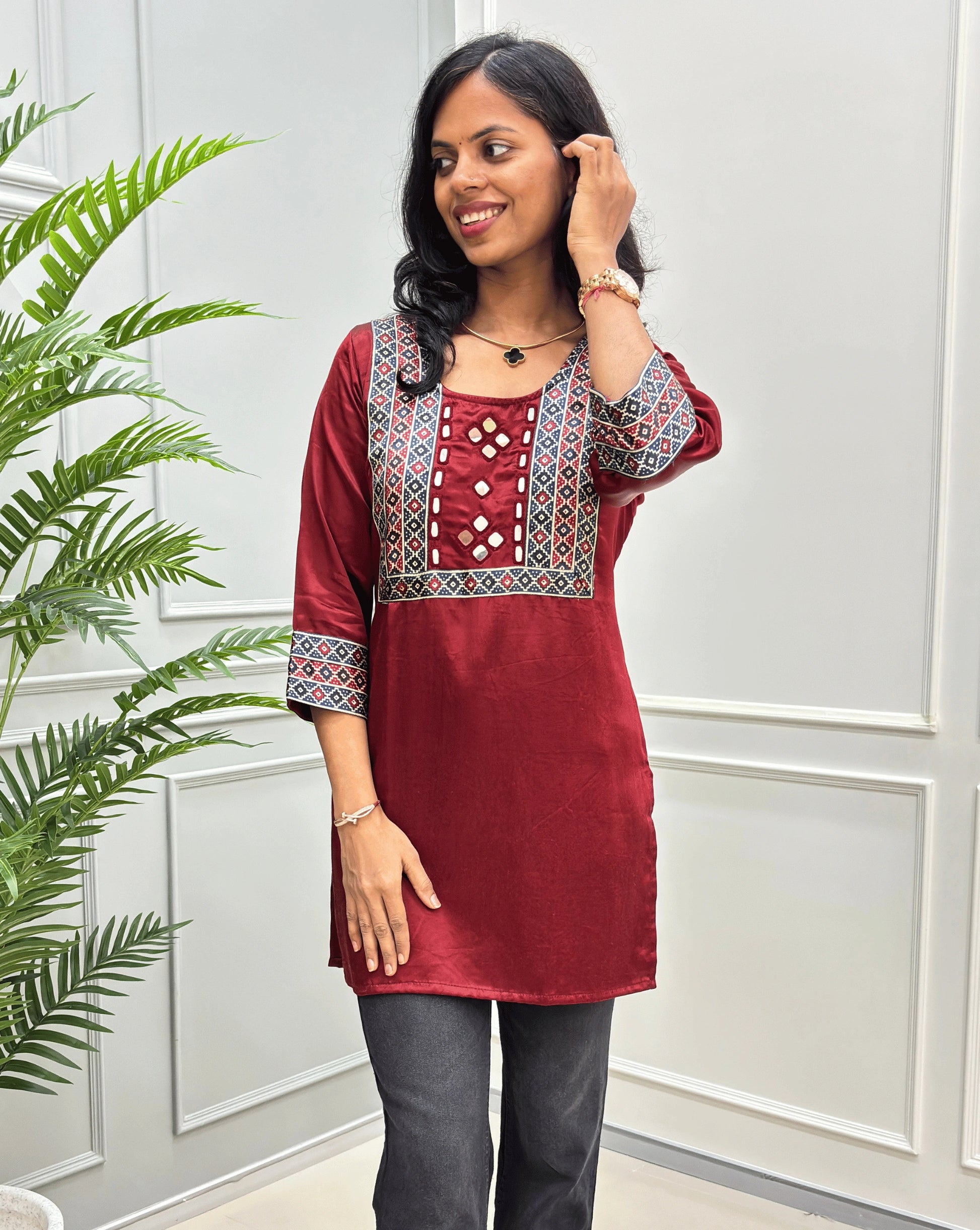 Close-up of mashru silk Red short kurti  with intricate multicolor geometric embroidery on the neckline and sleeves, paired with dark gray pants, standing in front of a white paneled wall with a green plant on the left.