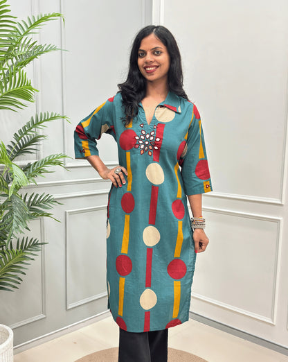 Front-view of Teal Green Women's Ajrakh Vegetable dyed plant based Cotton Long-Kurti with a geometric pattern of Red, beige, and yellow circles, Hand-mirror work on front in flower shape & Sleeves cuffs,  paired with black Flared pant, standing in a room with a potted plant and paneled walls.
