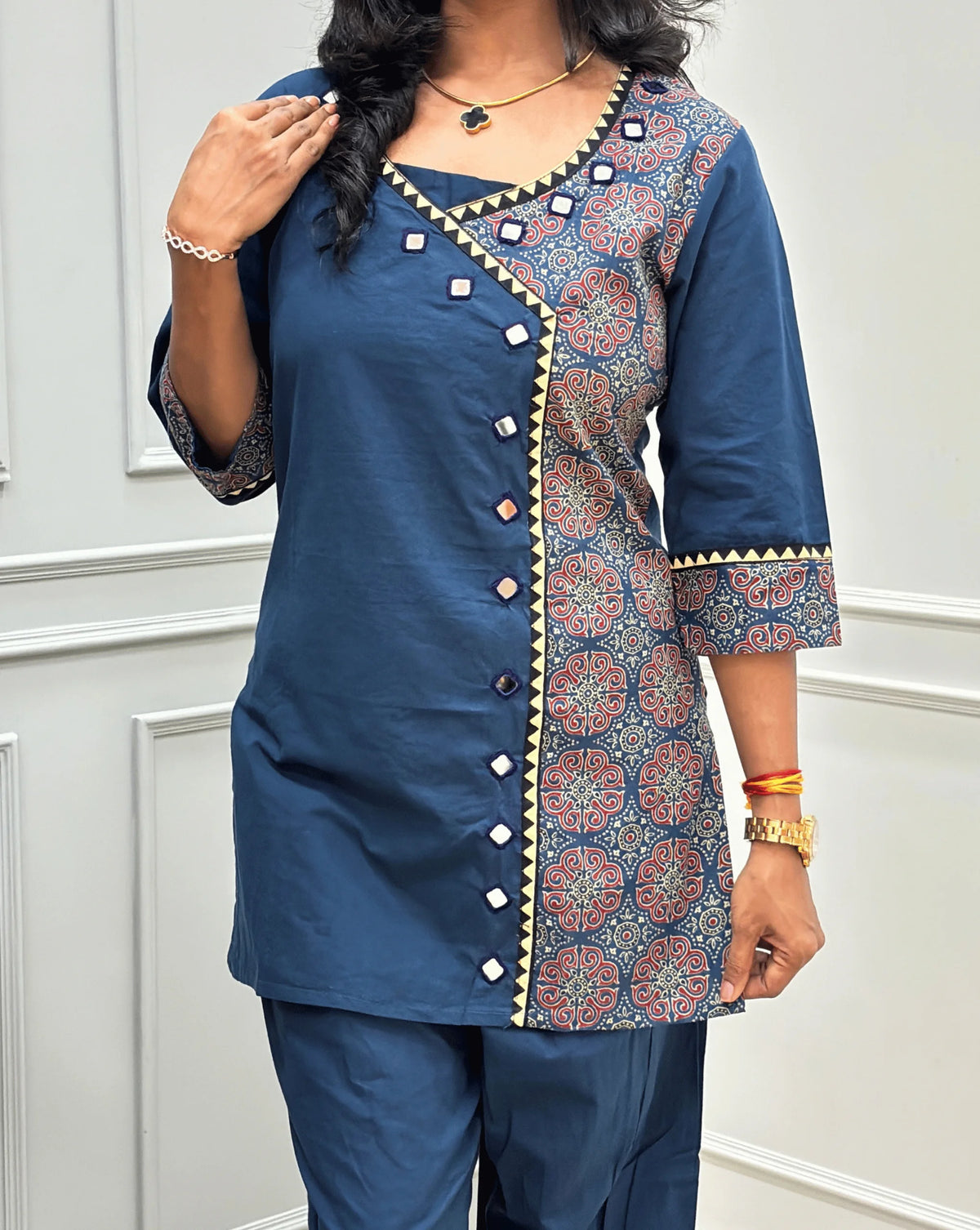 Close-up view of Blue Women's Ajrakh Pure Cotton 2-peice Co-Ord set Featured with V-shaped neck with a Ajrakh printed panel on the front & Sleeves, Angrakha Patterned on front, Real Hand-Mirror work on front Along with 2 side pockets in pant, standing on a round woven rug in a modern room with white paneled walls.