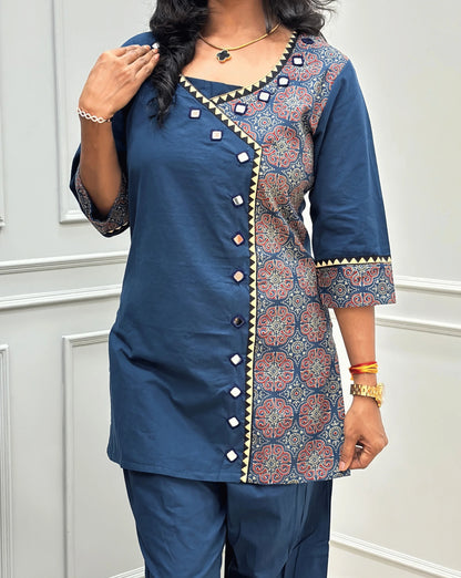 Close-up view of Blue Women's Ajrakh Pure Cotton 2-peice Co-Ord set Featured with V-shaped neck with a Ajrakh printed panel on the front & Sleeves, Angrakha Patterned on front, Real Hand-Mirror work on front Along with 2 side pockets in pant, standing on a round woven rug in a modern room with white paneled walls.