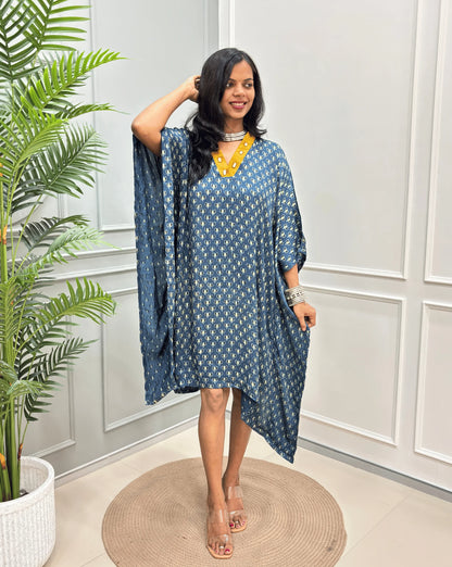 Blue Women's Ajrakh Pure Modal Silk Knee Length Kaftan with a black Floral print , standing on a round woven rug in front of a white paneled wall next to a potted plant.