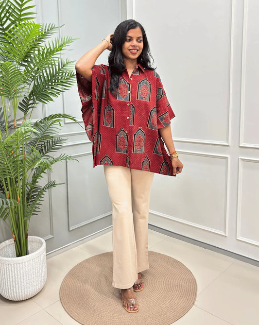 Red Women's Ajrakh Pure Cotton Short Kaftan Shirt with a Black, Grey & Red Floral Block Print, paired with beige flared pants, standing on a round woven rug in front of a white paneled wall with a potted plant.