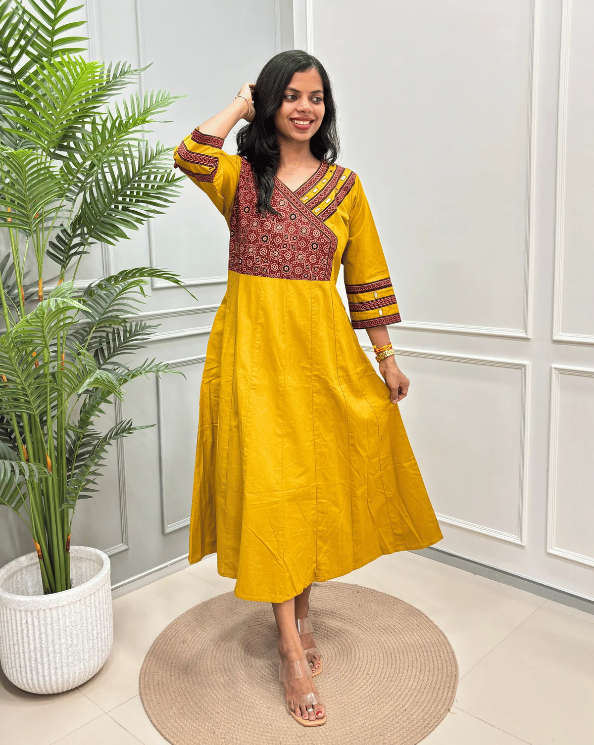Yellow Women's Ajrakh Pure Cotton A-line Anarkali with a Red base & Black floral yoke patterned on Front & sleeve cuffs, standing on a round woven rug in front of a light gray paneled wall.