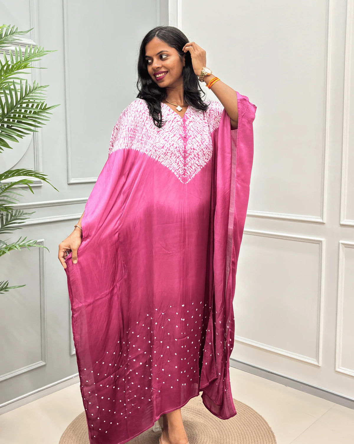 Front-Side View of Pink Women's Ajrakh Pure Modal Silk Shibori Kaftan Featured with Long-length, with a 3 shades of Pink with ajrakh print on chest & Bandhani Small dots with white on Bottom, standing on a round woven rug in front of a white paneled wall.