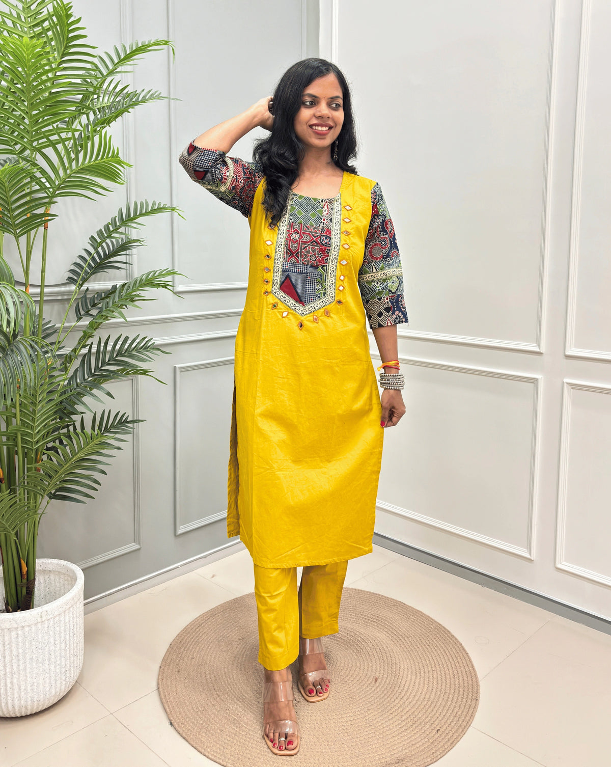 Yellow women's Ajrakh Vegetable dyed plant based Cotton CoOrd set with Multi-colour ajrakh Print detailed embroidered on neckline & Sleevess with beige border on work ,  paired with matching teal pants, standing on a round woven mat in a bright room with a potted plant.