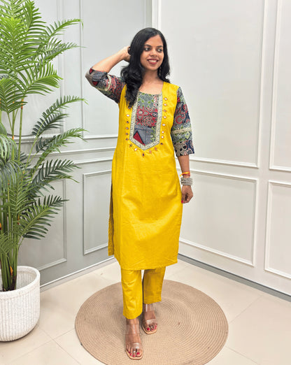 Yellow women's Ajrakh Vegetable dyed plant based Cotton CoOrd set with Multi-colour ajrakh Print detailed embroidered on neckline & Sleevess with beige border on work ,  paired with matching teal pants, standing on a round woven mat in a bright room with a potted plant.
