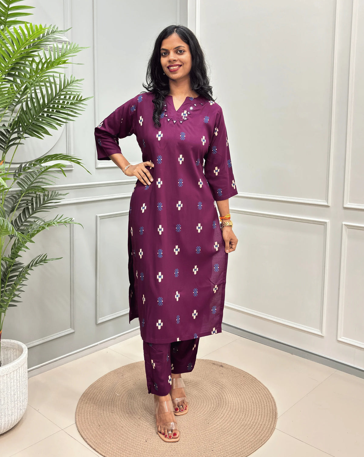 Purple Women's Ajrakh Vegetable Dyed Cotton Long Co-Ord Set with white and blue geometric Print, along with Same Matching pant, standing on a round woven mat in front of a white paneled wall with a green plant on the left.