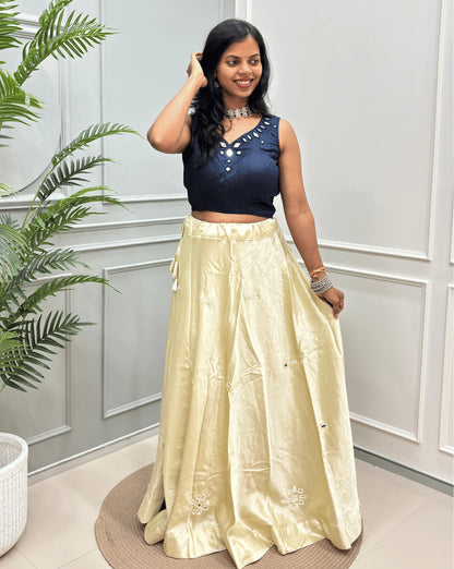Ivory Women's Pure Mashru silk  two-piece traditional outfit consisting of a sleeveless  Blue crop top with Real Hand mirror work and a Ivory flared skirt with embroidered details, set against a white paneled wall with a green plant on the left.