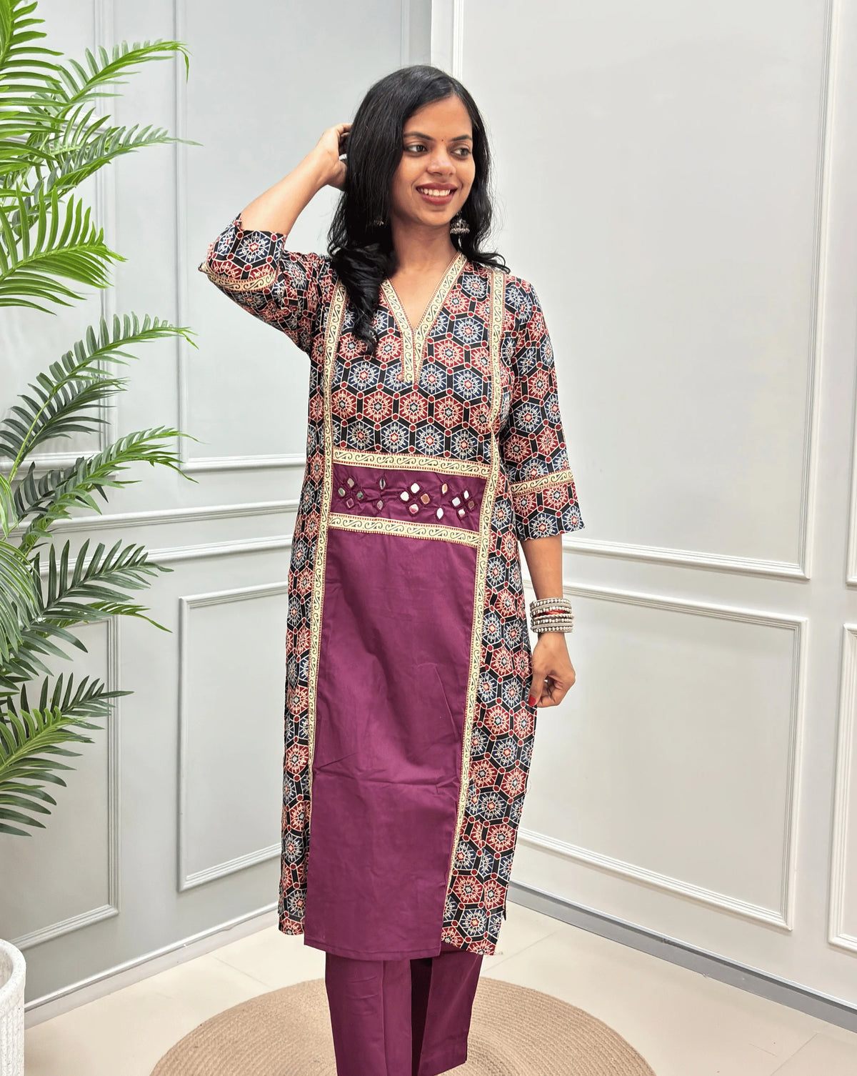 Front-view of Purple Women's Ajrakh pure cotton 2 peice Co-Ord set with a black base & an abstract of blue & Red Geometric print on Front Chest to Side & Sleeves , Hand-mirror work on Front as Belt, standing on a round woven mat in front of a white paneled wall with a potted plant.