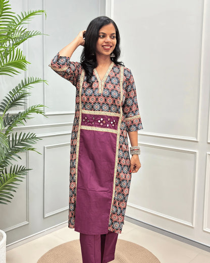 Front-view of Purple Women's Ajrakh pure cotton 2 peice Co-Ord set with a black base & an abstract of blue & Red Geometric print on Front Chest to Side & Sleeves , Hand-mirror work on Front as Belt, standing on a round woven mat in front of a white paneled wall with a potted plant.