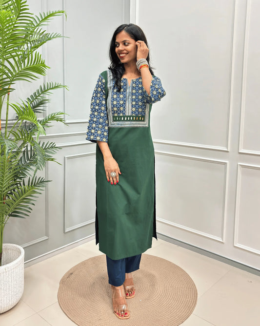 Dark Green Women's Hand-block Ajrakh Pure Cotton Long Kurti with a blue base , an abstract of black & beige Floral Print yoke on front & Full sleeves, paired with blue Pant, standing on a round woven mat in front of a white paneled wall.