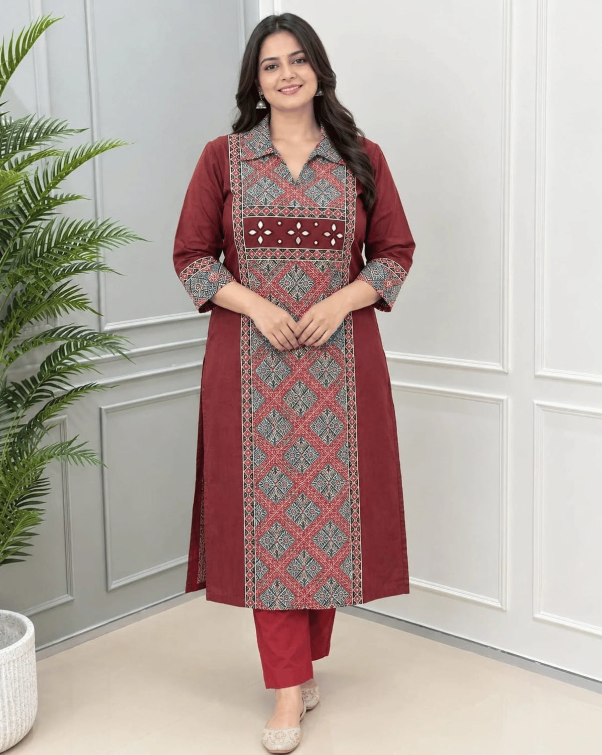 Maroon women's Ajrakh Vegetable Dyed Plant Based Cotton Long-Kurti with a red Base, an Abstract of Black  & Beige geometric patterned panel on front & Sleeves Cuffs, paired with red pants, standing in a bright room with a white & Grey paneled wall ,on left side a potted plant.