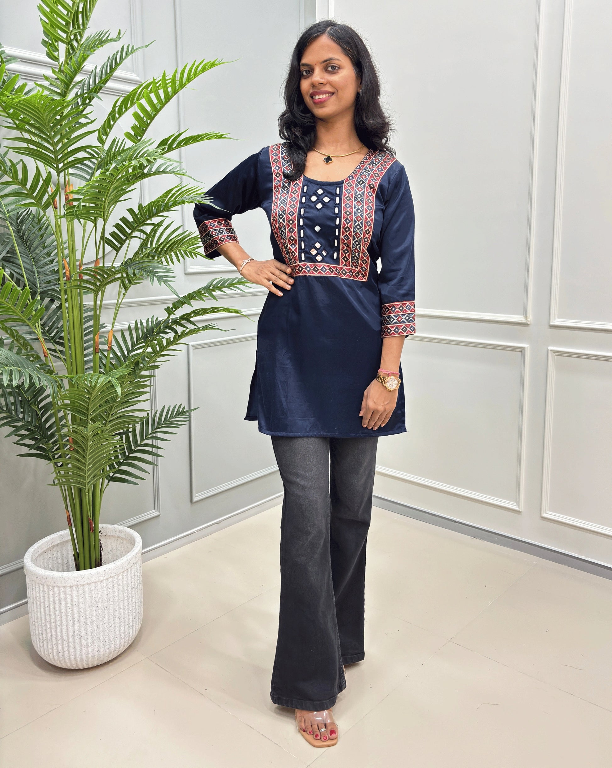 navy blue kurta with red and gold embroidered details, paired with dark gray flare jeans, standing in a room with a potted plant.