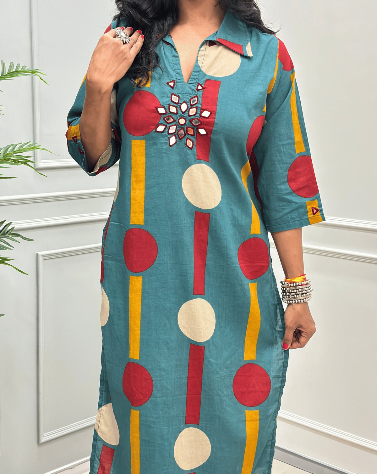 Close-up of Teal Green Women's Ajrakh Vegetable dyed plant based Cotton Long-Kurti Featured Collar V-neck & Three-quarter Sleeves with a geometric pattern of Red , beige, and yellow circles, Hand-mirror work on front in flower shape & Sleeves cuffs,  paired with black Flared pant, standing in a room with a potted plant and paneled walls.