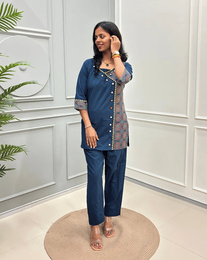 Blue Women's Ajrakh Pure Cotton 2-peice Co-Ord set with a Ajrakh printed panel on the front & Sleeves, Angrakha Patterned on front, standing on a round woven rug in a modern room with white paneled walls.