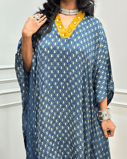 Close-up view of Blue Women's Ajrakh Pure Modal Silk Knee Length Kaftan with a black Floral print, with Contrast yellow Colour V-neck, along with Real Hand-Mirror embroidery, standing on a round woven rug in front of a white paneled wall next to a potted plant. 